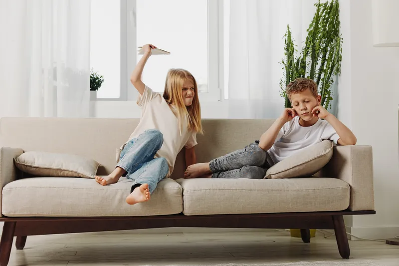 Two children sitting on beige sofa in bright living room, girl holding paper plane above boy while he covers ears. Concept of sibling interaction, playfulness, and childhood moments.