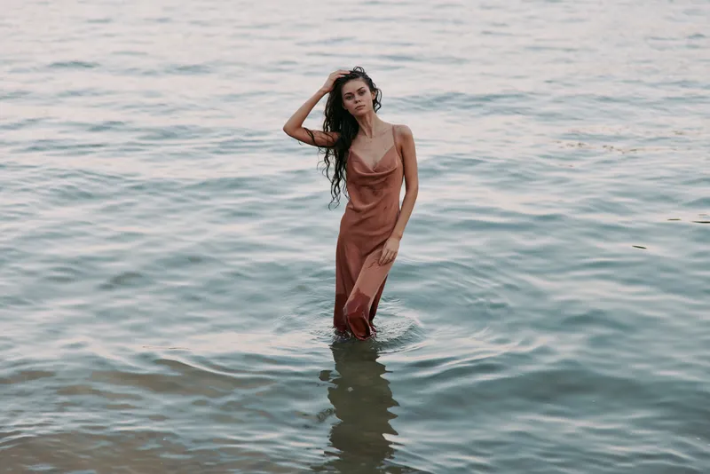 A woman wades through shallow sea water in a satin slip dress, posing gracefully as gentle waves ripple around her, sunlight glinting on the surface, serene mood.