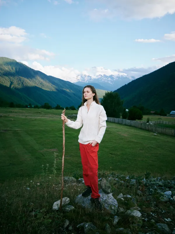 Woman stands on rocky ground in a white sweater and red pants, holding a wooden staff, amid a wide valley and mountain landscape under a blue sky