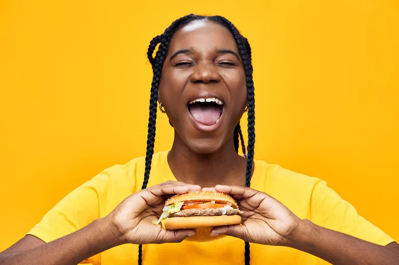 Happy young woman enjoying a delicious burger against a vibrant yellow background, showcasing joy and a love for fast food