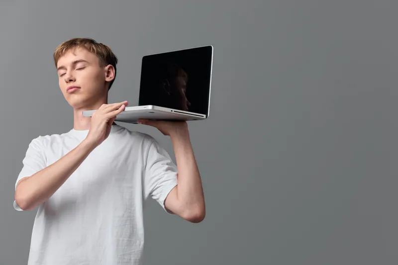Young man balancing a laptop on his shoulder eyes closed calm expression casual white t shirt minimal studio setting conceptual moment of portable tech and focus