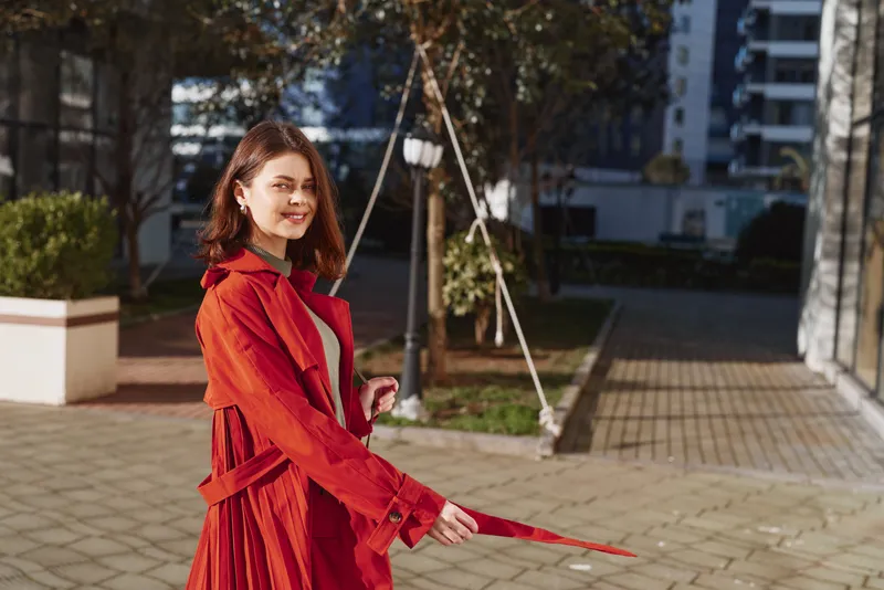 Smiling woman in a red coat holding a red umbrella in an urban courtyard, street style fashion, confident pose, warm daylight, city backdrop.