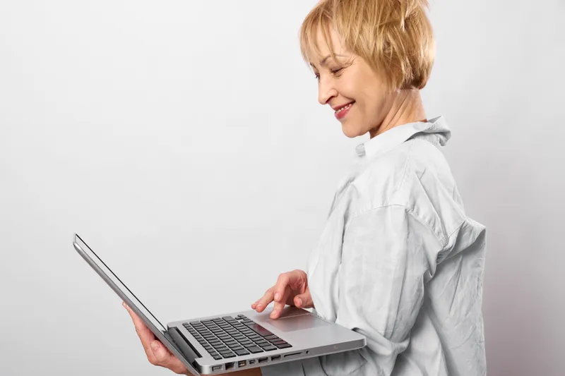 Woman with laptop, smiling office portrait, side profile, hands typing on keyboard, using technology, casual shirt, bright white background, indoor, soft light, calm
