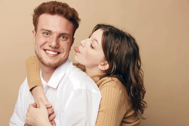 Happy young couple embraces and smiles in casual clothes on beige background. Woman hugs man from behind and playfully sticks out tongue. Concept of love, friendship, and joy.