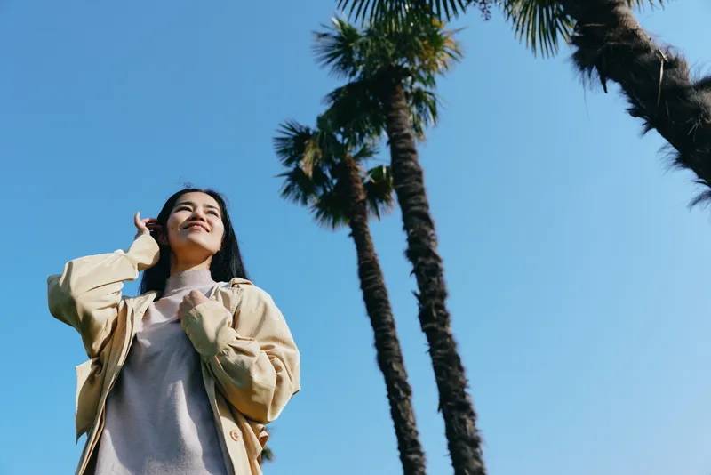 Woman posing under tall palm trees against a bright blue sky, enjoying sunshine in a casual jacket, relaxed mood, outdoor scenery, carefree fashion, confident stance, summer vibe