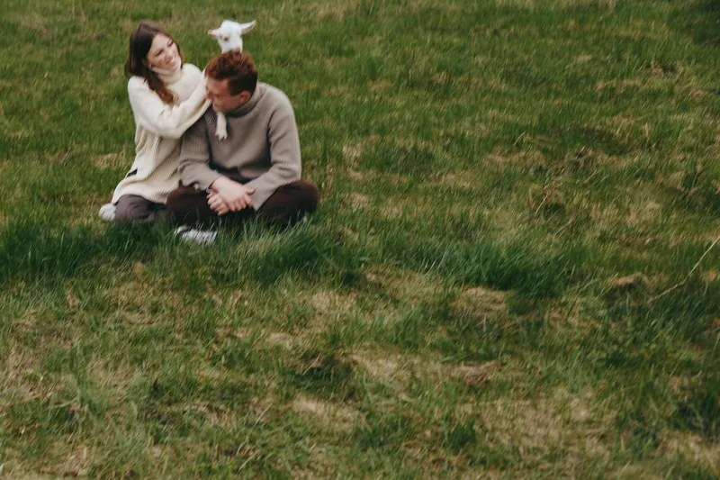 Couple sitting on a grassy field with a playful goat kid perched on the man's shoulder. Outdoor lifestyle moment showing affection, companionship, and rural life with warm, natural light.
