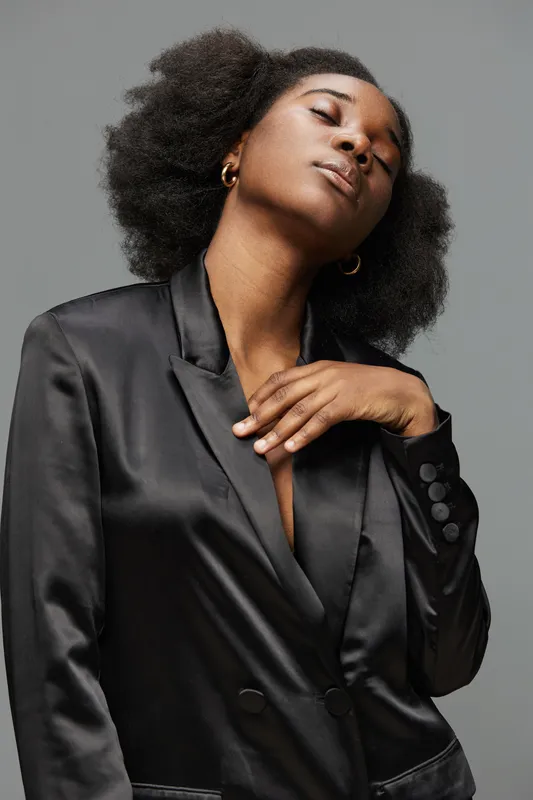 Confident Black woman in a sleek black satin blazer, exuding elegance and self assuredness against a neutral gray background, showcasing contemporary fashion