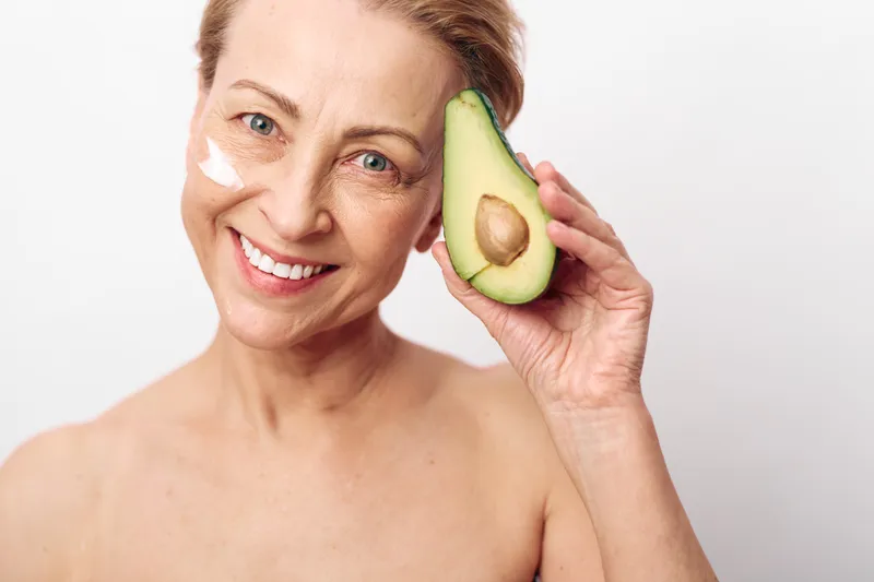 Smiling mature woman with natural skin holds avocado and applies cream on cheek. Concept of healthy skincare, natural beauty, wellness, and organic facial care for middle aged women.