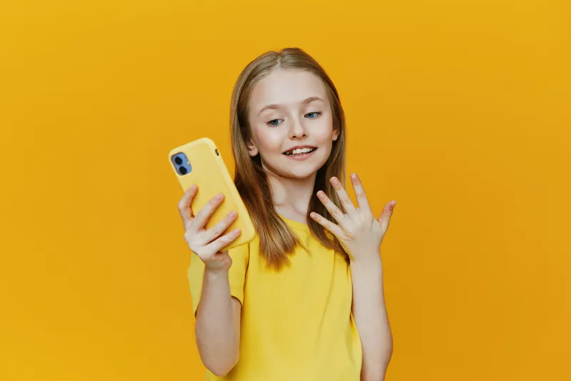 Happy girl in a yellow shirt taking a selfie with a smartphone against a bright yellow background, capturing joyful moments and youthful energy
