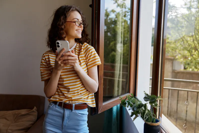 Young woman with curly hair in a striped yellow tshirt stands by an open window, smiling as she checks her phone in a cozy home setting with soft natural light, a plant nearby, and a relaxed mood