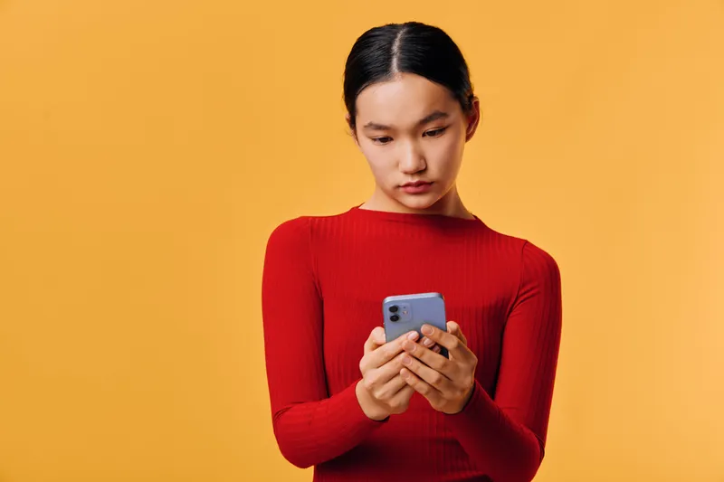 Young woman in red sweater looking at smartphone with focused expression indoors, isolated on orange background. Casual portrait of female person using mobile device for communication or browsing.