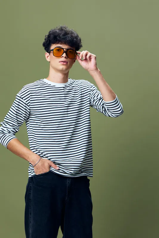 Young man wearing striped shirt and orange sunglasses posing confidently against green background, displaying casual style and relaxed attitude with hand in pocket.