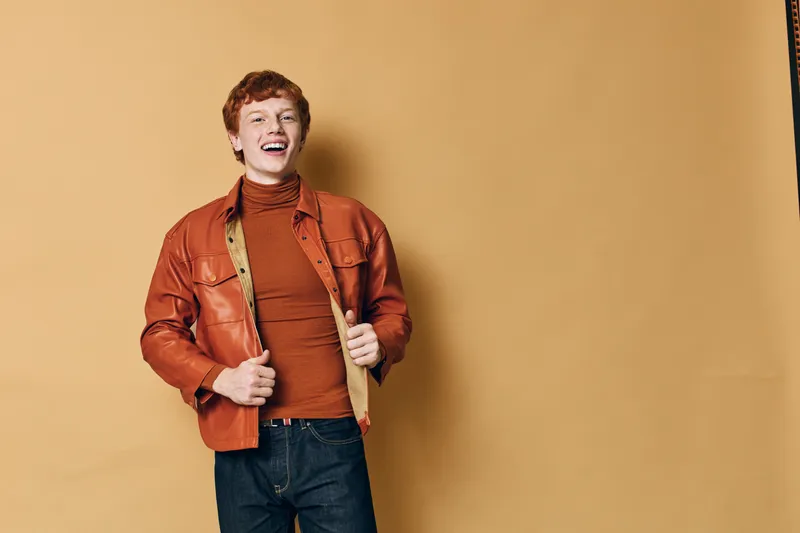 Young man with red hair wearing orange leather jacket and turtleneck smiling with hands in pockets against solid beige background indoor portrait