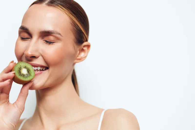 Woman kiwi fruit skincare beauty healthy fresh smile closeup Young woman holds kiwi by cheek for natural facecare, clean studio white background, joyful mood