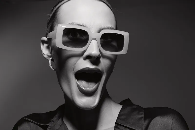 Stylish caucasian woman with pale skin wearing large sunglasses and hoop earrings makes surprised face in black and white studio portrait.