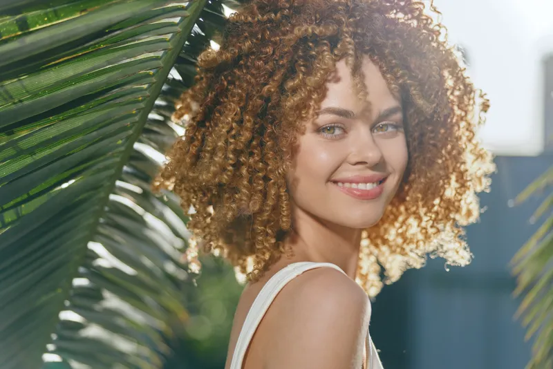 smiling woman with curly hair enjoying warm sunlight outdoors surrounded by green palm leaves in a bright summer day with natural beauty and happiness.