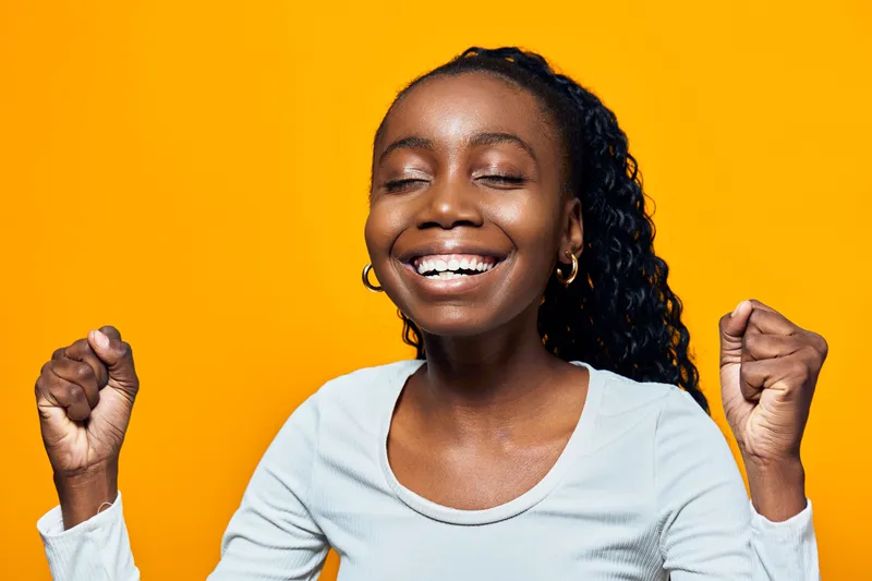 Joyful young Black woman celebrating success with a bright smile against a vibrant yellow background, wearing a light colored top and expressing happiness