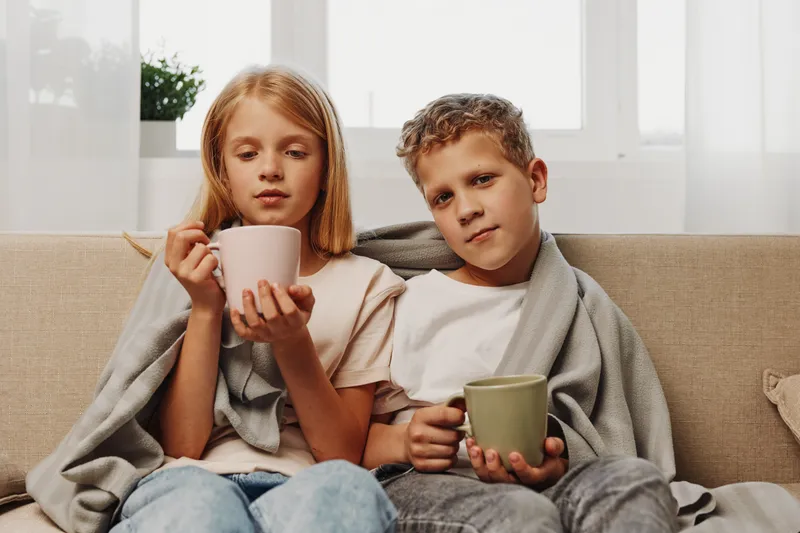 Two children wrapped in gray blankets sit on a beige sofa holding mugs, appearing cozy and relaxed in a bright room with large windows behind them.