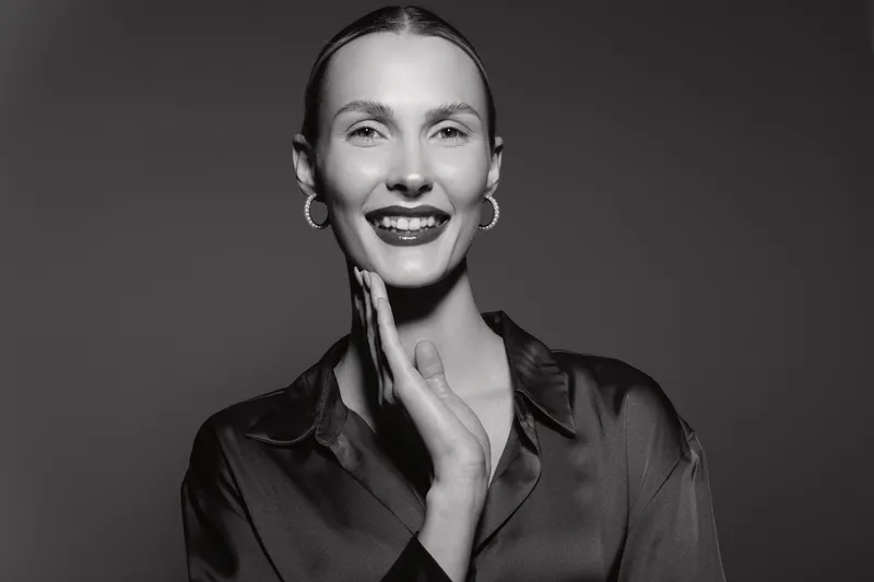 A smiling Caucasian woman with fair skin and slicked-back hair poses confidently in a satin blouse against a dark background. Concept of elegance and beauty.