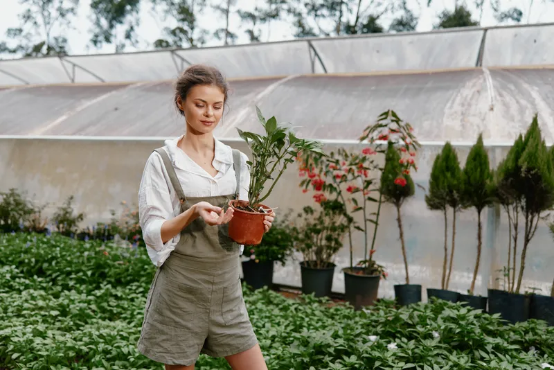 Young woman in a greenhouse holding a potted plant, exhibiting joy and connection with nature, dressed in casual green overalls, surrounded by greenery