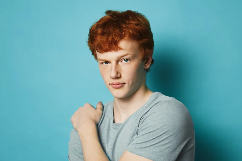 Young man with red hair and fair skin wearing gray t shirt on blue background looking confident and relaxed with casual pose and natural lighting in studio portrait.