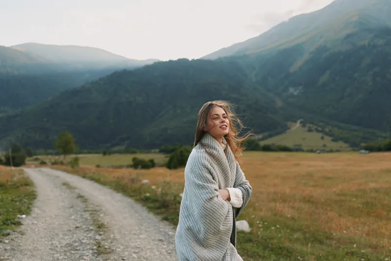 A woman wrapped in a cozy blanket stands on a rural path surrounded by green fields and mountains. She enjoys the fresh air and peaceful nature, embodying a relaxed lifestyle.