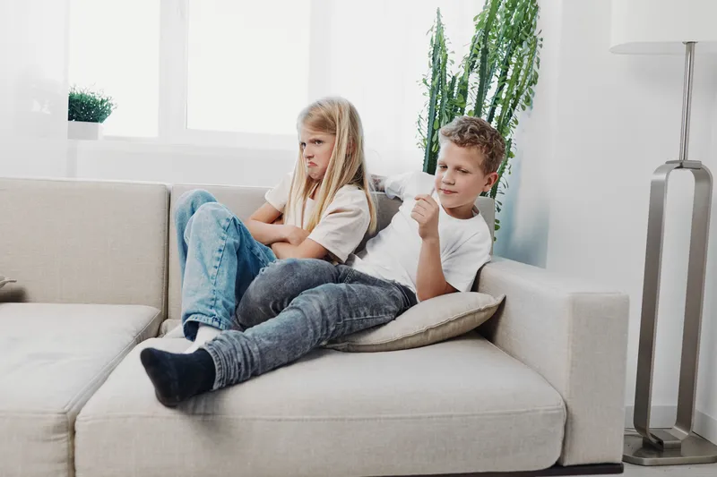 Upset girl and relaxed boy sitting apart on couch in bright living room. Concept of sibling conflict, family tension, and childhood emotions indoors.