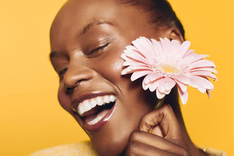 Close-up of happy African woman with dark skin holding pink flower near face on yellow background. Concept of joy, beauty, skincare, natural glow, and positive emotions in studio lighting.