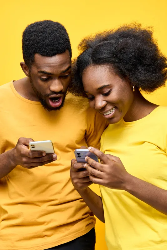 A joyful African couple interacting with their smartphones against a vibrant yellow backdrop, wearing matching yellow t shirts, showcasing excitement and connection