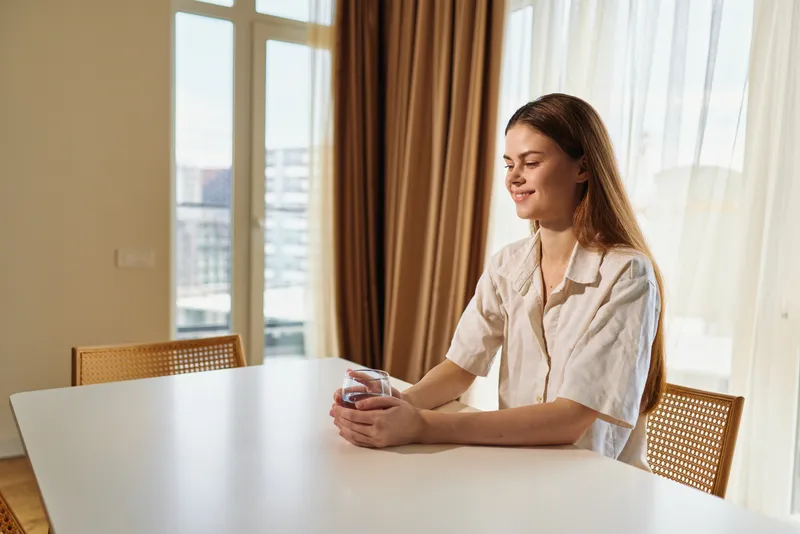 Woman portrait holding coffee at table in home morning beside window, smiling and relaxed in cozy interior with sunlight, casual outfit and peaceful domestic atmosphere.