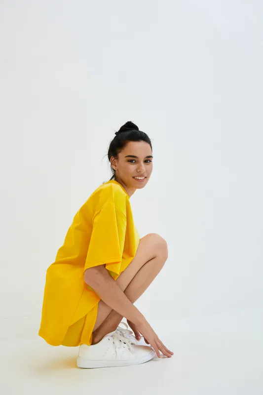 Young woman squatting in vibrant yellow dress and white sneakers, smiling confidently against a minimalistic white background with natural lighting and casual style.