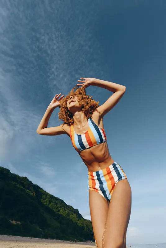 Confident woman in striped swimsuit on a beach, summer outdoors pose under bright blue sky, sunlit water and green hills. Vertical portrait, carefree vacation.