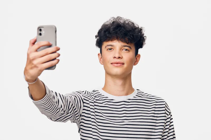 young man taking selfie with smartphone wearing striped shirt against white background, curly hair, casual style, youthful expression, digital lifestyle