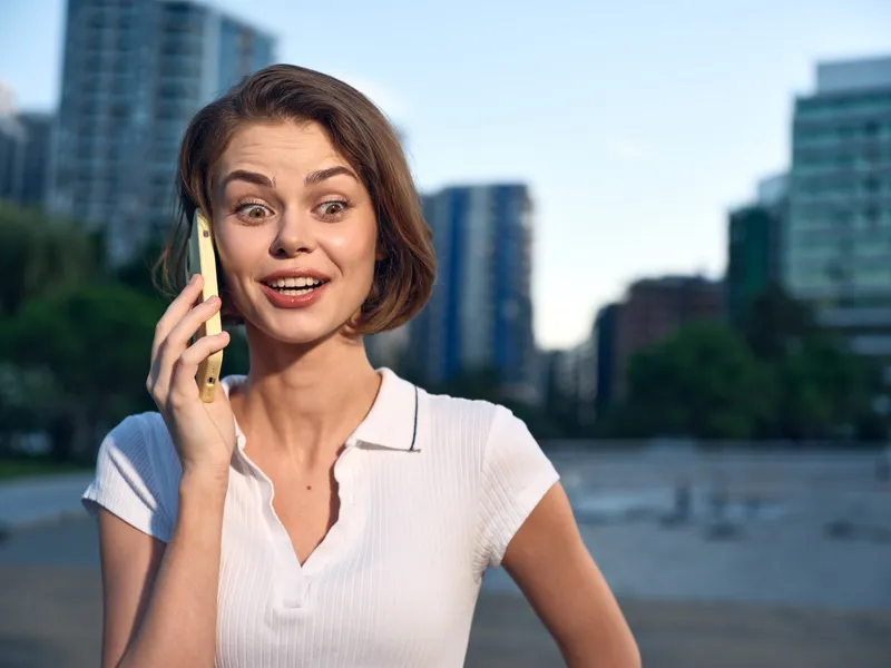 A cheerful professional woman speaks on a smartphone outdoors in an urban park setting. She smiles warmly, present and confident, with modern buildings in the background.