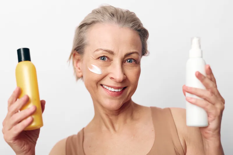 Smiling mature woman holds two skincare bottles with cream on cheek, posing on white background. Concept of beauty routine, facial care, anti aging, and healthy skin lifestyle.