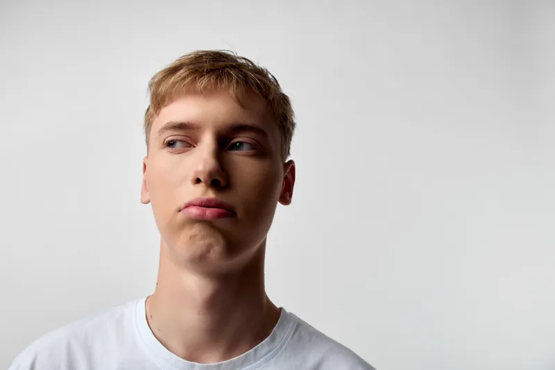 Young man with a puzzled expression and a pout, looking to the side in a white tshirt, studio setting. Subtle lighting highlights a casual mood, thoughtful vibe, and contemporary style.
