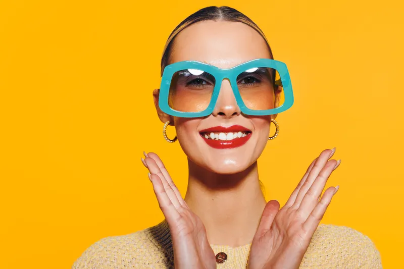 Smiling woman with large blue sunglasses and red lipstick poses against bright yellow background. Fashion portrait with cheerful mood and trendy accessories in studio lighting.