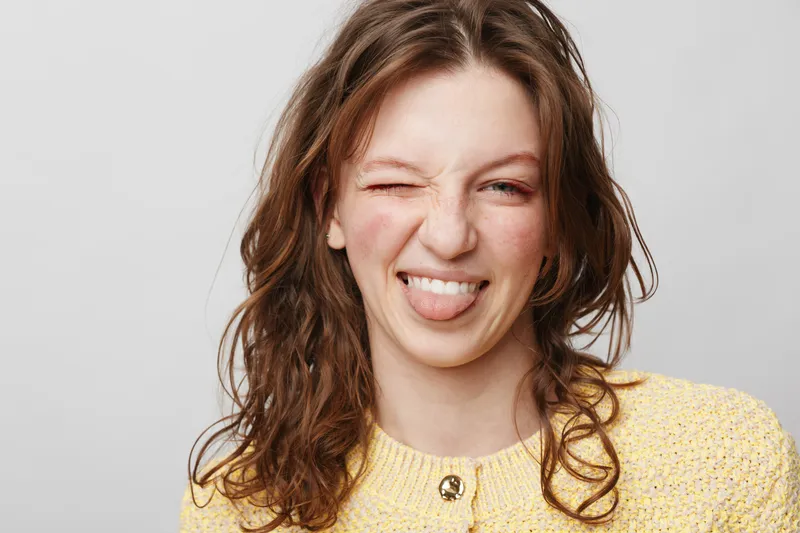 Young woman with curly brown hair wearing yellow sweater makes playful face sticking out tongue and winking. Studio portrait on light grey background with casual and fun mood.