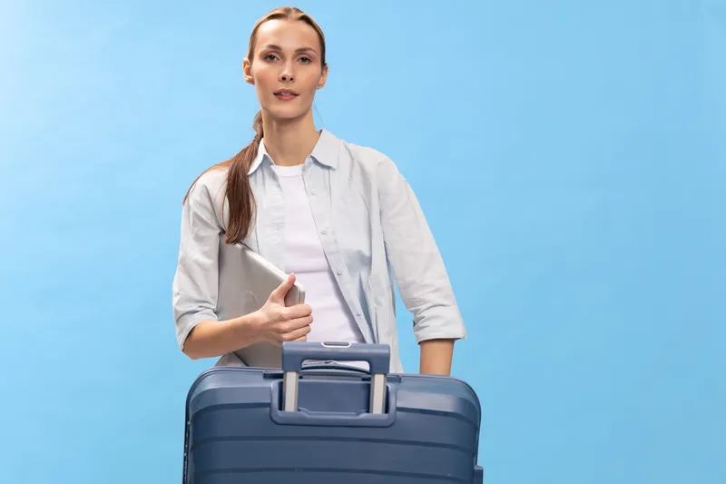 Young woman holding laptop stands behind blue suitcase on blue background. Business travel concept with casual outfit and confident facial expression, preparing for trip or journey.