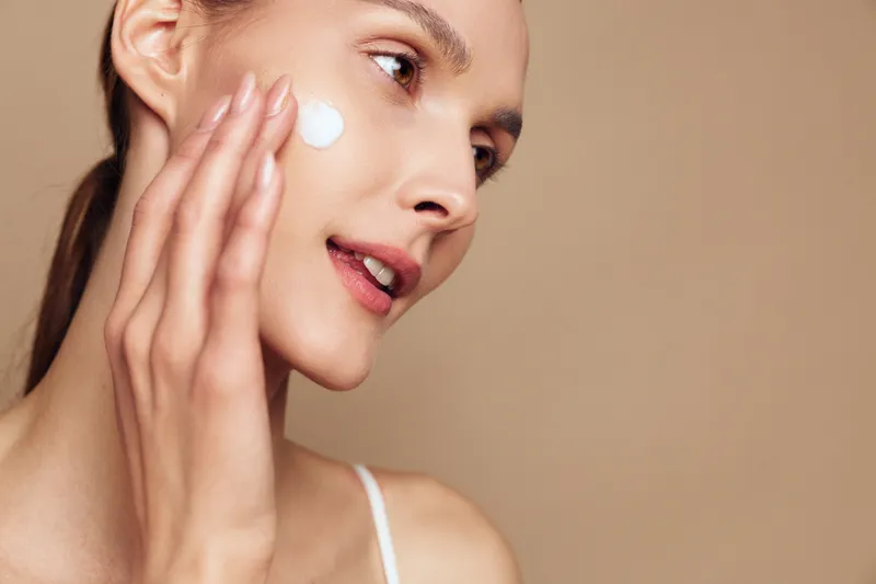 Woman applying cream to her cheek in a closeup portrait, soft light, skincare moment, smooth skin, gentle touch, natural tones, calm mood, minimal background