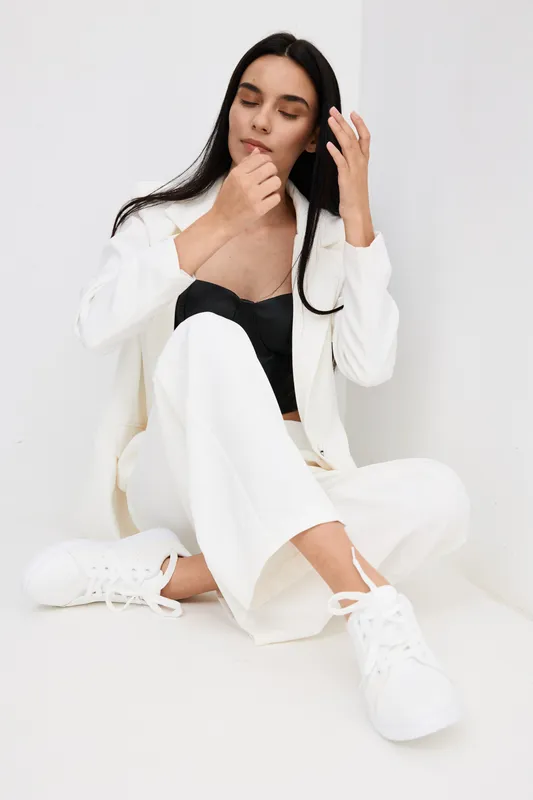 Relaxed woman with long dark hair dressed in stylish white suit and black top sitting on floor against white background in bright minimalistic setting.