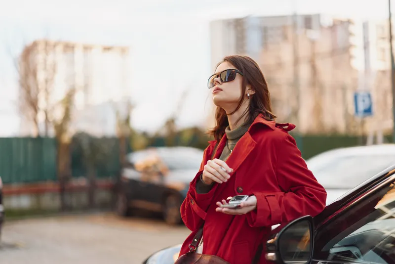 Confident woman in a red coat wearing sunglasses, holding a camera on a city street. Street style fashion, urban vibe, chic and ready for a photo shoot today.