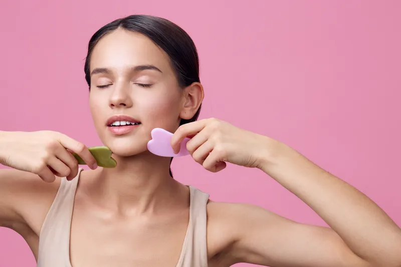 Young woman using gua sha tools for facial massage on a pink background, promoting beauty and relaxation, with soft expressions and natural makeup