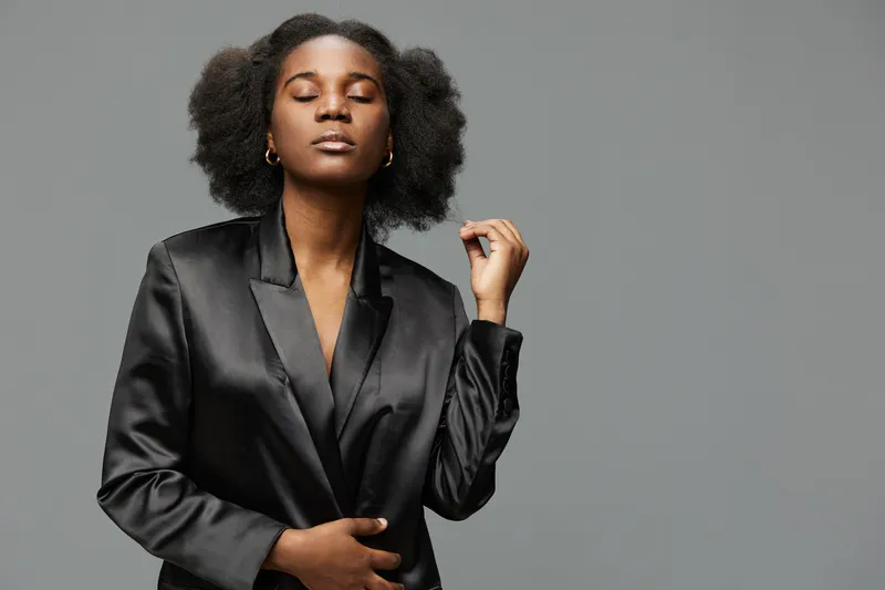 Confident young black woman in black blazer with natural hair, closed eyes, and serene expression against a gray background, portraying empowerment and style