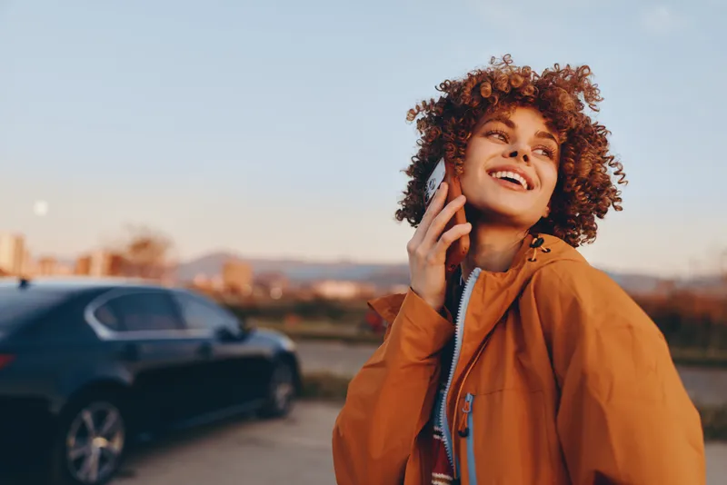 Woman lifestyle portrait outdoors, curly hair, smiling while talking on phone, warm sunset light, casual jacket, street view with parked car softly blurred behind, relaxed confident mood