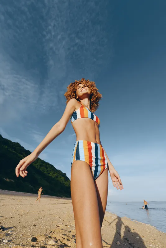 Woman beach striped swimsuit summer ocean blue sky sunlight curly hair, relaxed standing on sand, gentle waves, distant green mountains, bright midday sun, natural colors, outdoor vacation mood