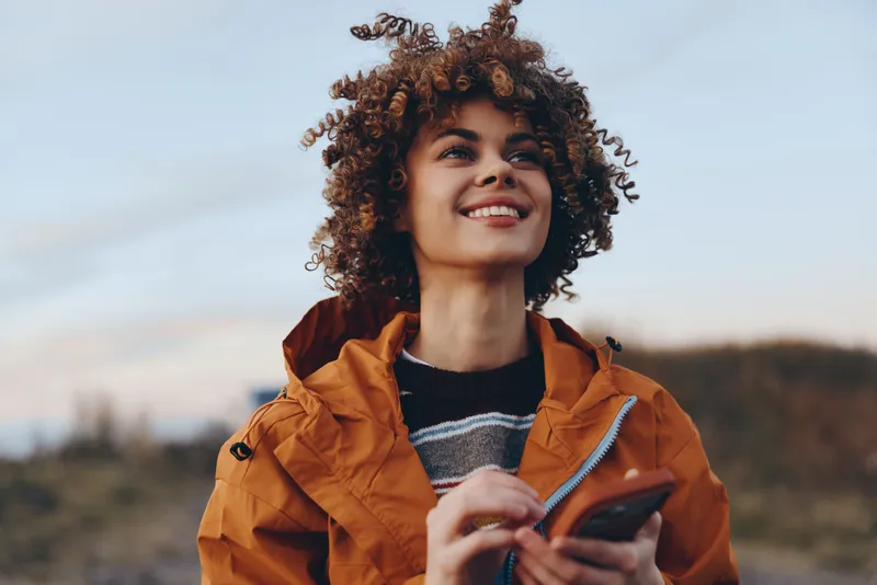 Woman lifestyle portrait outdoors, smiling with curly hair, orange jacket, holding smartphone in hands, sunny sky and rural landscape behind, casual travel mood, close up today.
