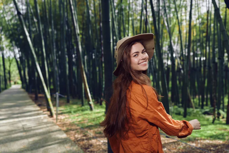 Woman in hat and orange sweater walking on a paved path through bamboo forest, smiling over shoulder. Outdoor portrait, travel lifestyle and relaxed happy nature walk scene.