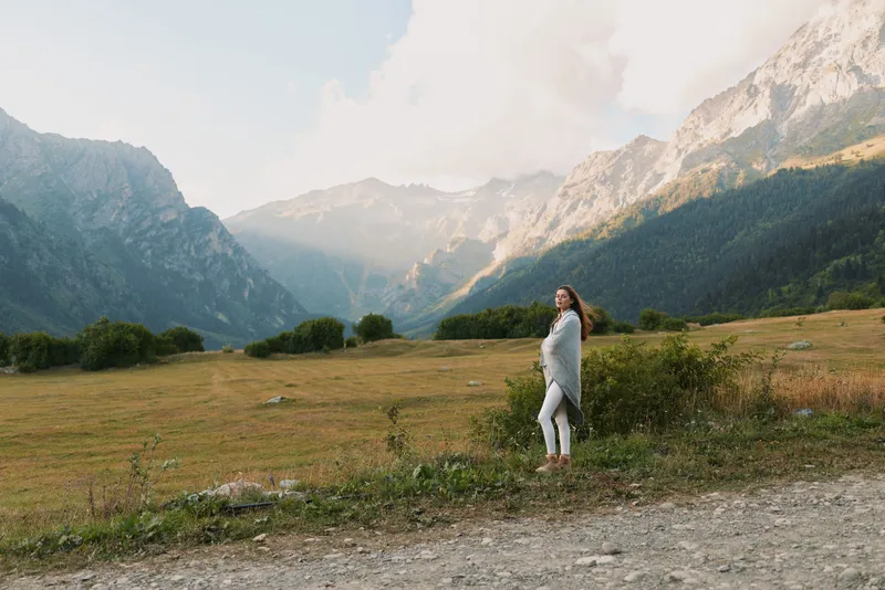 Casual woman outdoors in a mountain valley, standing and gazing into distance, soft light, natural colors, peaceful mood, expansive landscape backdrop with distant peaks