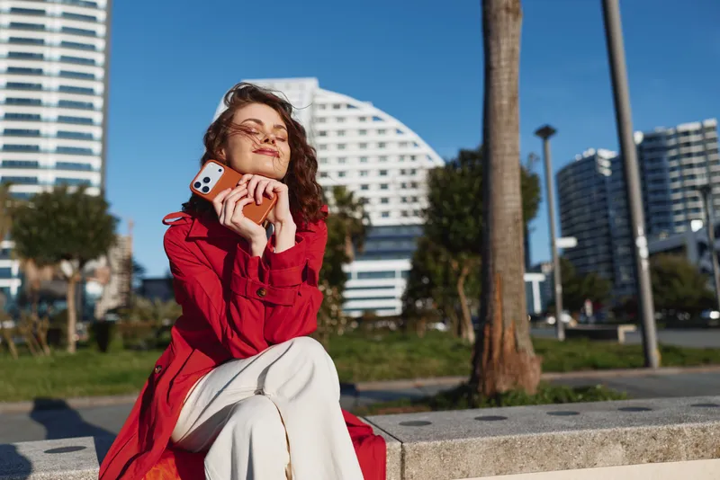 Young woman in a red coat enjoys sunlight while sitting outdoors holding a smartphone near her face with closed eyes and a peaceful expression.